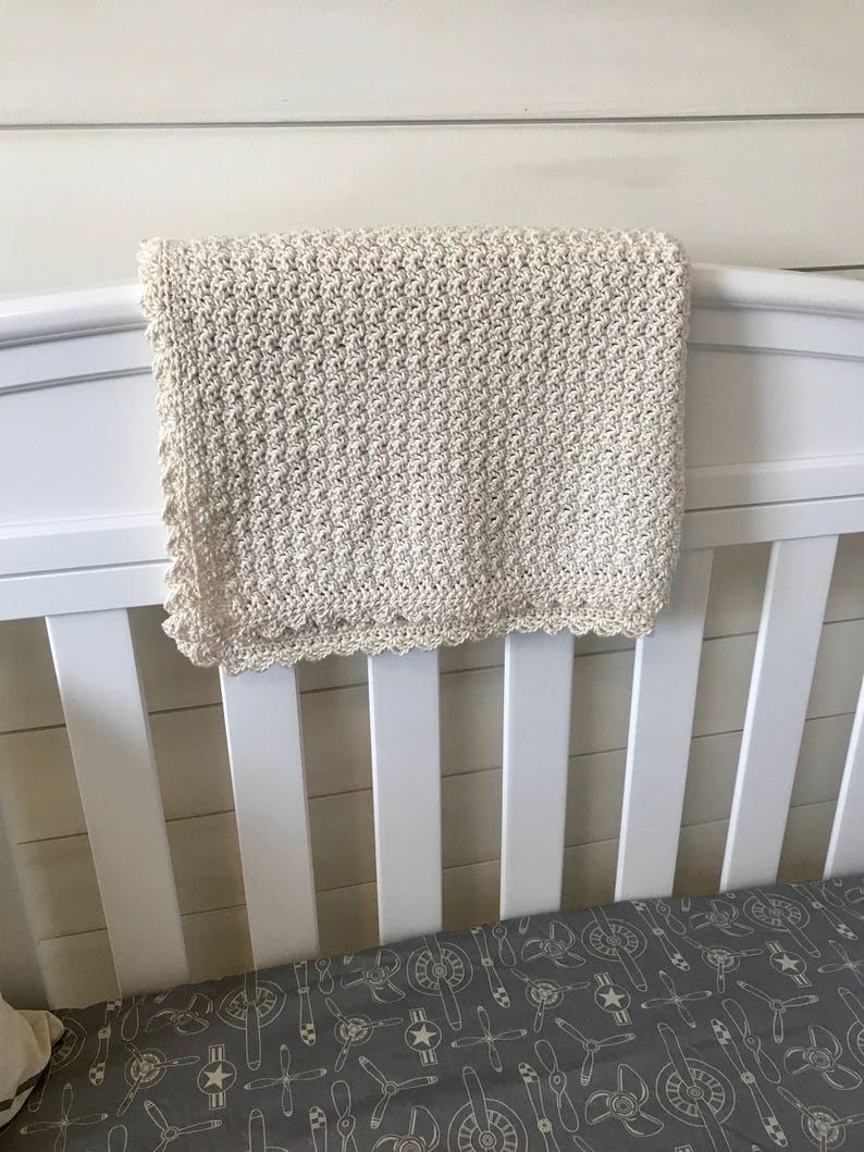 May include: A white crocheted baby blanket with a scalloped edge hangs on a white crib. The crib has a gray and white patterned sheet with a propeller design.