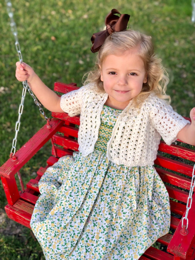 May include: A young girl wearing a white crocheted cardigan over a floral dress sits on a red wooden swing. She has a brown bow in her hair and is smiling at the camera.