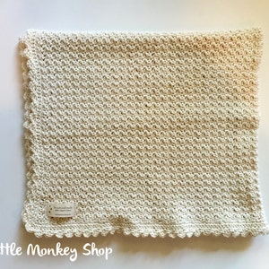May include: A cream-colored crocheted baby blanket with a scalloped edge. The blanket is made with a textured stitch pattern. The label reads "For the little ones of the world, with love, 100% organic cotton, littlemonkeyshop.com".