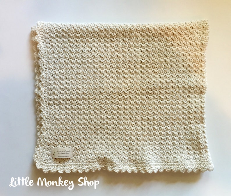 May include: A cream-colored crocheted baby blanket with a scalloped edge. The blanket is made with a textured stitch pattern. The label reads "For the little ones of the world, with love, 100% organic cotton, littlemonkeyshop.com".