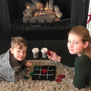 May include: Two children play a game of tic-tac-toe on a black and white knitted board with red and green game pieces. They are sitting on a fluffy beige rug in front of a fireplace with a lit fire.