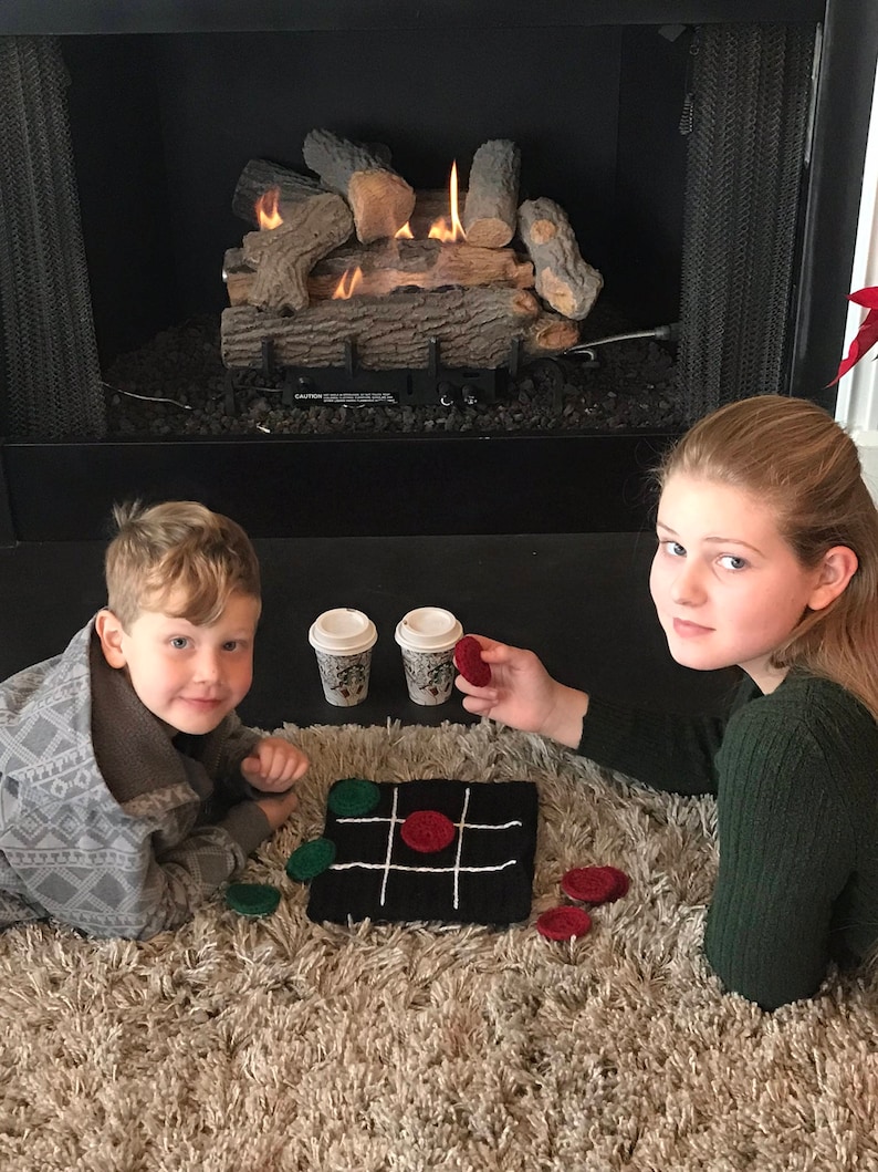 May include: Two children play a game of tic-tac-toe on a black and white knitted board with red and green game pieces. They are sitting on a fluffy beige rug in front of a fireplace with a lit fire.