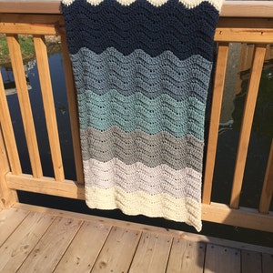 May include: A crocheted blanket with a wavy pattern in shades of blue, gray, and white. The blanket is hanging on a wooden railing.