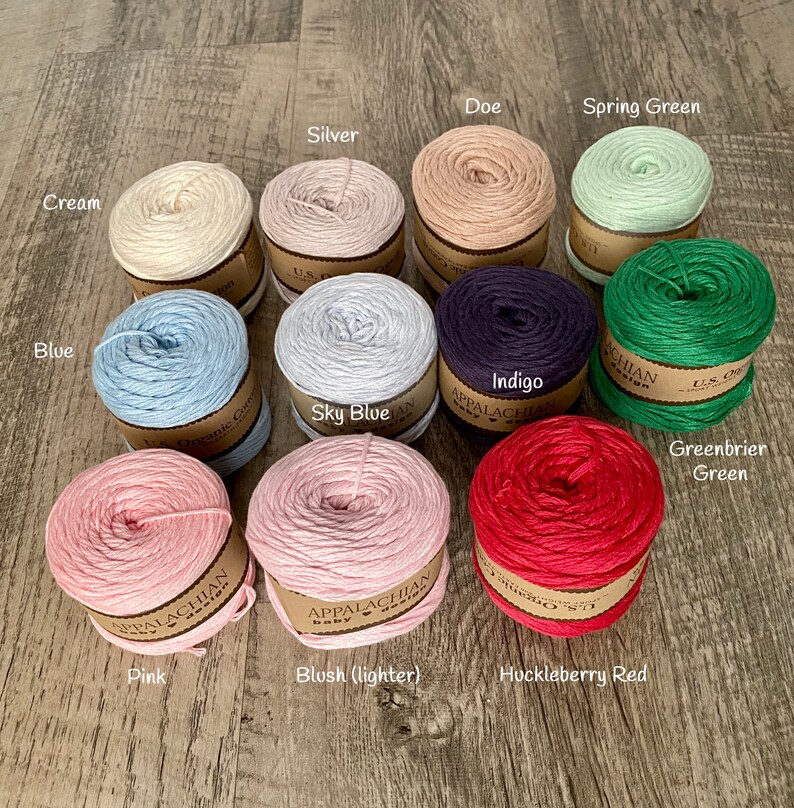 May include: Assortment of 12 skeins of yarn in various colors, including cream, blue, sky blue, indigo, greenbrier green, spring green, silver, doe, pink, blush (lighter), and huckleberry red. Each skein is labeled with the yarn color and "Appalachian Baby Yarn".