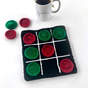 May include: A black crocheted tic-tac-toe game board with white lines and red and green crocheted game pieces. There are extra game pieces on the table next to the board.