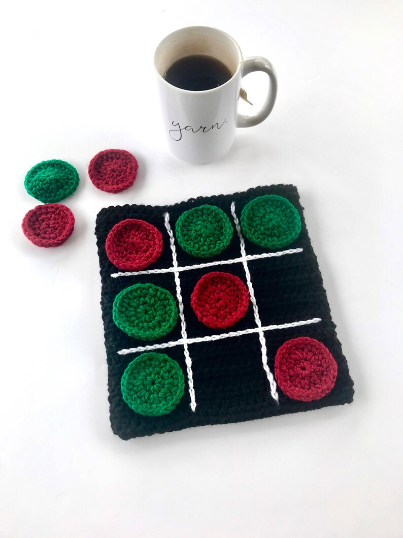 May include: A black crocheted tic-tac-toe game board with white lines and red and green crocheted game pieces. There are extra game pieces on the table next to the board.