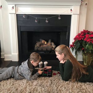 May include: Two children play a game of tic-tac-toe on a brown rug in front of a fireplace. The fireplace has a white mantel with a string of Christmas lights. A poinsettia plant is in a terracotta pot to the right of the fireplace.