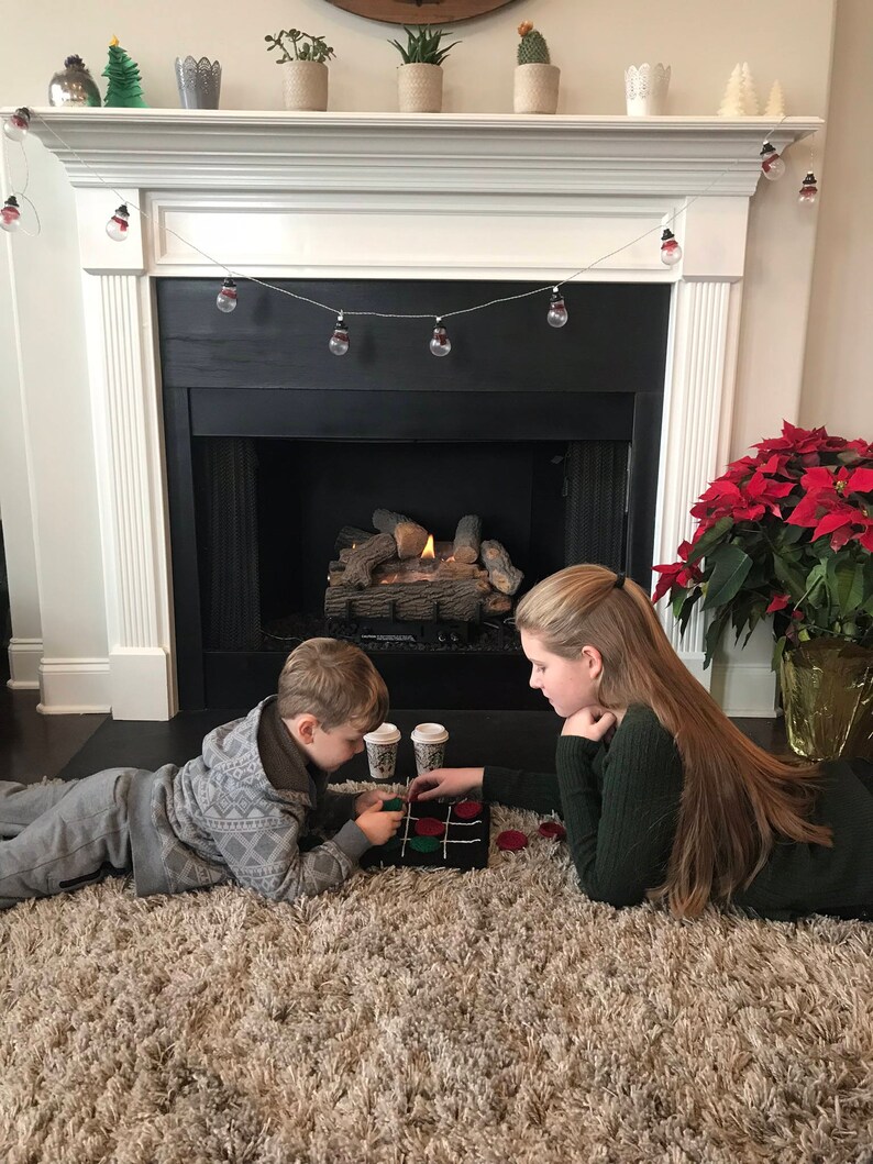 May include: Two children play a game of tic-tac-toe on a brown rug in front of a fireplace. The fireplace has a white mantel with a string of Christmas lights. A poinsettia plant is in a terracotta pot to the right of the fireplace.