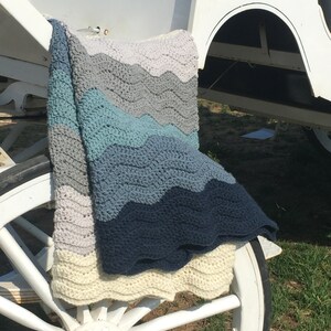 May include: A crocheted blanket with a wavy pattern in shades of blue, gray, and white. The blanket is draped over a white wagon wheel.