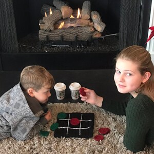 May include: Two children play a game of tic-tac-toe on a crocheted board with red and green game pieces. They are sitting on a fluffy rug in front of a fireplace with a faux log set.