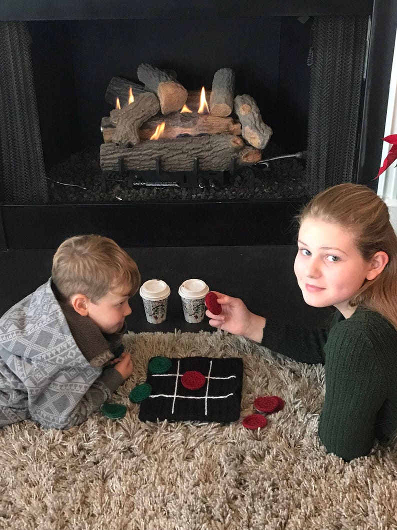 May include: Two children play a game of tic-tac-toe on a crocheted board with red and green game pieces. They are sitting on a fluffy rug in front of a fireplace with a faux log set.