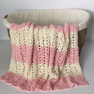 May include: A crocheted baby blanket with pink and cream stripes. The blanket is folded and draped over a wicker basket.