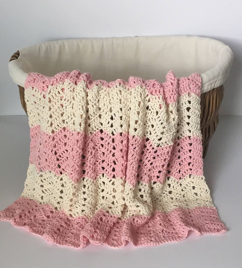 May include: A crocheted baby blanket with pink and cream stripes. The blanket is folded and draped over a wicker basket.