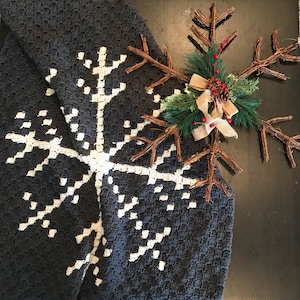 May include: A dark gray crocheted blanket with a large white snowflake design. A decorative twig snowflake with greenery, red berries, and a burlap bow rests on the blanket, creating a winter theme.