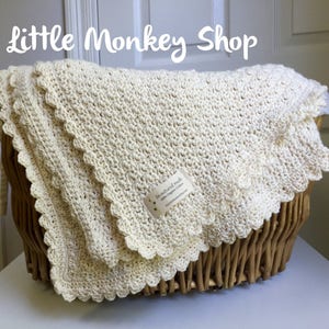 May include: A cream-colored crocheted baby blanket with a scalloped edge, folded and displayed in a wicker basket. The blanket is made with natural materials and has a tag that reads "In natural made with Organic Cotton Little Dragon Creations".