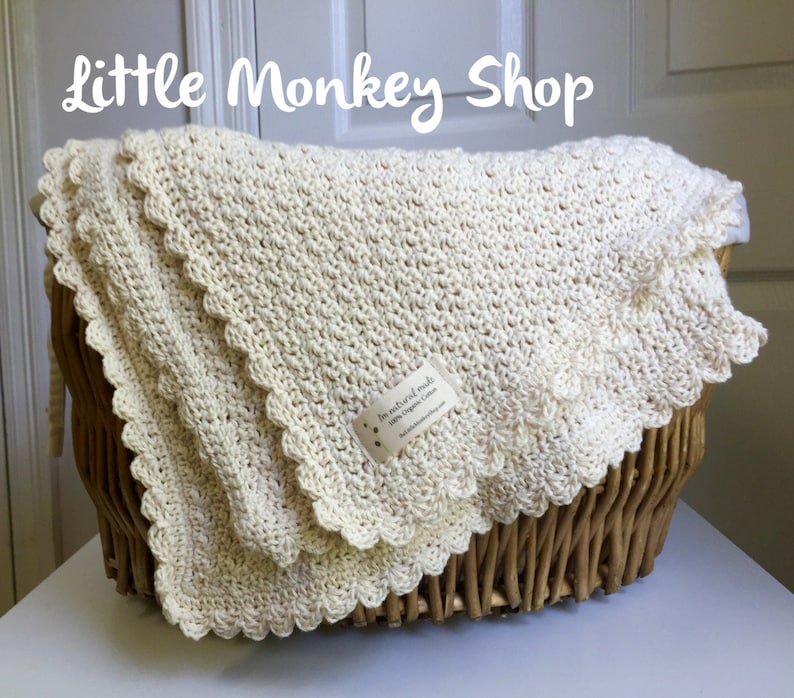 May include: A cream-colored crocheted baby blanket with a scalloped edge, folded and displayed in a wicker basket. The blanket is made with natural materials and has a tag that reads "In natural made with Organic Cotton Little Dragon Creations".