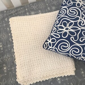 May include: A white crocheted baby blanket with a scalloped edge, laying on a gray crib sheet with a white airplane pattern.