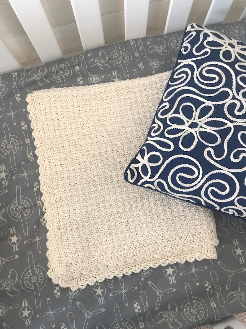 May include: A white crocheted baby blanket with a scalloped edge, laying on a gray crib sheet with a white airplane pattern.