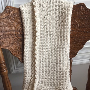 May include: A cream-colored crocheted baby blanket with a scalloped edge. The blanket is draped over a wooden chair.
