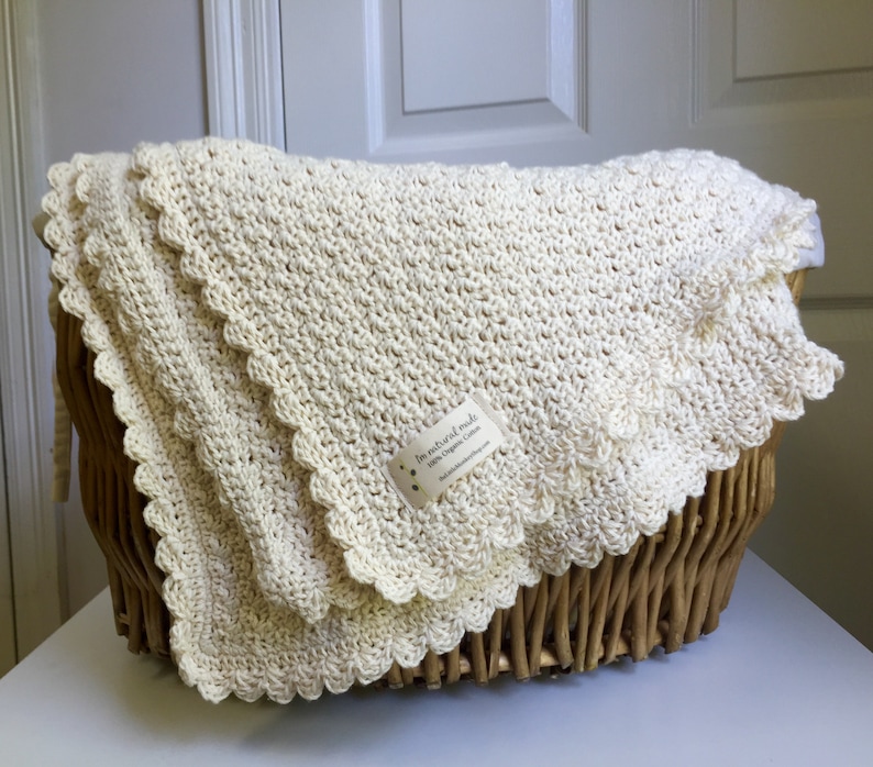 May include: A cream-colored crocheted baby blanket with a scalloped edge. The blanket is folded and placed in a wicker basket. A small tag with the text "In natural nude 100% Organic Cotton" is attached to the blanket.