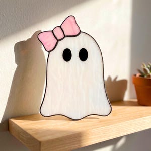 Peut inclure: Un ornement fantôme en vitrail composé d'un corps blanc semi-translucide avec un bord inférieur ondulé. Le fantôme présente deux yeux circulaires noirs opaques et un nœud en verre rose clair fixé sur le côté supérieur gauche de sa tête.