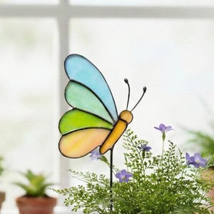 May include: A colorful butterfly garden stake with blue, green, and yellow stained glass wings. The butterfly has a yellow body and black antennae. It is placed in a pot of green plants with purple flowers.