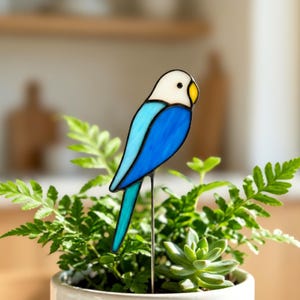 Puede incluir: Un adorno de jardín con forma de pájaro de cristal. Tiene la cabeza blanca, el cuerpo azul y el pico amarillo. Montado sobre una estaca de metal, se coloca en una maceta con plantas verdes. Decoración de jardín.