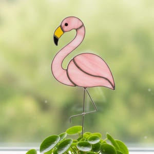 Könnte beinhalten: Ein rosafarbener Flamingo aus Glas mit gelbem Schnabel und schwarzen Akzenten. Der Flamingo steht auf einem dünnen Metallstab, ein Bein angehoben. Der Hintergrund ist ein verschwommenes Grün.