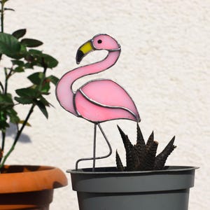 Könnte beinhalten: Rosa gefärbter Glas-Flamingoständer für den Garten mit gelbem Schnabel, der in einem grauen Topf mit einer Sukkulente steht.