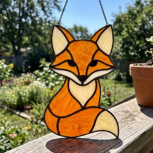 Puede incluir: Un atrapasoles de vidrieras con forma de zorro, que presenta piezas de vidrio de color naranja vibrante y crema unidas por líneas de soldadura de metal oscuro. El zorro se muestra en posición sentada con una cola grande y curva y orejas puntiagudas.