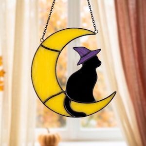 May include: A stained glass suncatcher shaped like a yellow crescent moon with a black cat silhouette wearing a purple pointed hat. The piece is constructed with dark metal lead lines and includes a black metal chain for hanging purposes.
