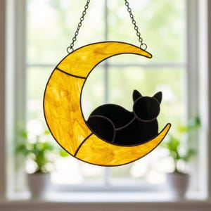 May include: A stained glass suncatcher featuring a crescent moon made of marbled yellow glass with a solid black glass cat silhouette resting on the curve. The piece is suspended by a thin metal chain attached to the top of the moon frame.