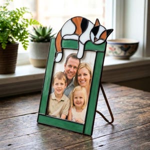 May include: A green stained glass picture frame with a family photo inside. The frame is decorated with a cat design in white, orange, and black. The frame is supported by a metal stand and sits on a wooden surface.