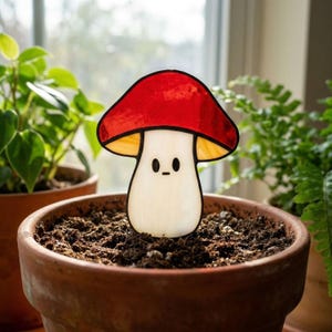 May include: A stained glass mushroom stake with a red cap, yellow gills, and a white stem. The mushroom has a cute face and is placed in a terracotta pot filled with soil. The background includes green plants and a window.
