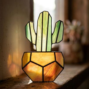 Op de afbeelding: Gebrandschilderd glazen cactus in een geometrische pot. De cactus is groen en geel, met zwarte contouren. De pot is geel, oranje en bruin. Het stuk wordt op een houten oppervlak gepresenteerd.