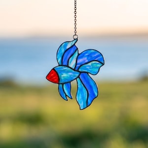 Könnte beinhalten: Ein Buntglas-Sonnenfänger in Form eines Kampffisches, bestehend aus irisierenden blauen und hellblauen Glassegmenten mit dunklen Metalllötlinien. Der Fisch hat einen Kopf aus rotem Glas und ist an einer dünnen Metallkette zum Aufhängen befestigt.