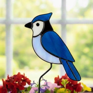 Puede incluir: Decoración de pájaro arrendajo azul de vidrieras con detalles en negro y vientre blanco. El pájaro está posado sobre un fino soporte de metal negro, con flores coloridas en primer plano. Las alas y el cuerpo del pájaro son de un azul vibrante.