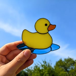 Può includere: Un acchiappasole in vetro colorato a forma di papera di gomma gialla con becco arancione, caratterizzato da un occhio nero e linee di saldatura scure. La papera è posizionata su una base in vetro blu che rappresenta l'acqua, contro un cielo.