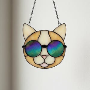 Op de afbeelding: Een gebrandschilderd glazen kattenvanger met een crème, oranje en witte kleurstelling. De kat draagt een ronde, zwart omrande zonnebril met iriserende glazen. Opgehangen aan een zwarte ketting, ideaal voor een raam.