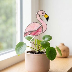 Puede incluir: Una estaca de planta de flamenco de vidrieras rosa con pico amarillo y punta negra se encuentra en una maceta rosa pequeña con plantas verdes. El flamenco tiene un marco de metal plateado y está cerca de una ventana.