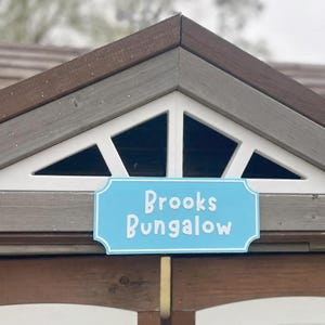 May include: A light blue sign with the words "Brooks Bungalow" in white lettering is mounted on a wooden structure. The sign is rectangular with a decorative border. The structure has a brown roof and white trim.