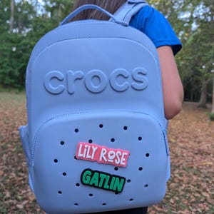 May include: A light blue backpack with a textured surface and the word crocs embossed on the top. The front pocket features multiple ventilation holes and two rectangular charms, one pink with the text LILY ROSE and one green with the text GATLIN.