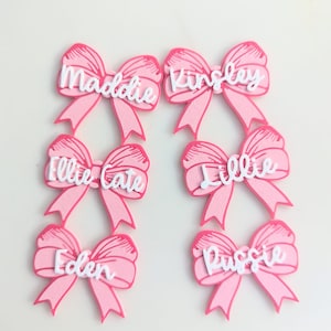 May include: Six pink bows with white lettering. The bows are decorated with the names Maddie, Kinsley, Ellie Cate, Lillie, Eden, and Pussie.