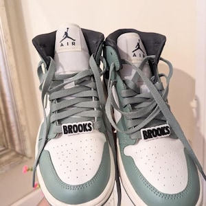 May include: A pair of white and sage green high-top sneakers with black laces and the word "BROOKS" on each shoe.