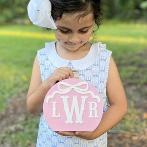 May include: A pink, round piggy bank with a white bow design and the letters "LWR" in white. The piggy bank is being held by a person wearing a blue and white dress with a white collar and a white bow in their hair.