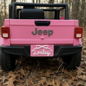 May include: A pink Jeep-branded ride-on toy car with black accents. The rear features a license plate that reads "Loxley" in a cursive font. The toy car is parked on a bed of fallen leaves.
