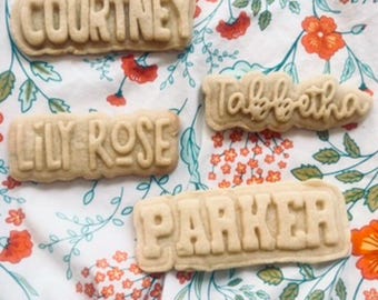 May include: Four light-colored cookies with names embossed on them: "Courtney", "Lily Rose", "Parker", and "Tabbetha". The cookies are on a floral patterned fabric with orange and red flowers.