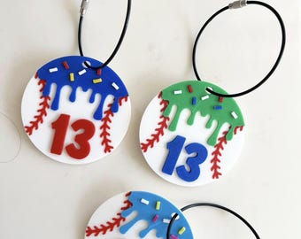 May include: Three baseball-themed keychains. Each keychain features a white circle with red baseball stitching, a number 13, and colorful dripped icing. The icing colors are blue, green, and red with sprinkles. Each keychain has a black wire loop.