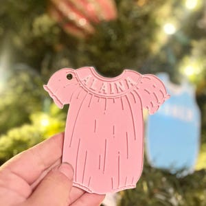 May include: A pink baby onesie Christmas tree ornament, with the name "ALAINA" engraved on the collar. The ornament is held in front of a Christmas tree, with a blue ornament visible in the background.