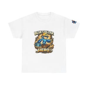 Op de afbeelding: Wit T-shirt met een cartoon illustratie van een blauw geklede bouwvakker met een gele helm. De afbeelding bevat de tekst "BLUE COLLAR HERO" in een gestileerd lettertype, tegen een achtergrond van gereedschap en tandwielen.